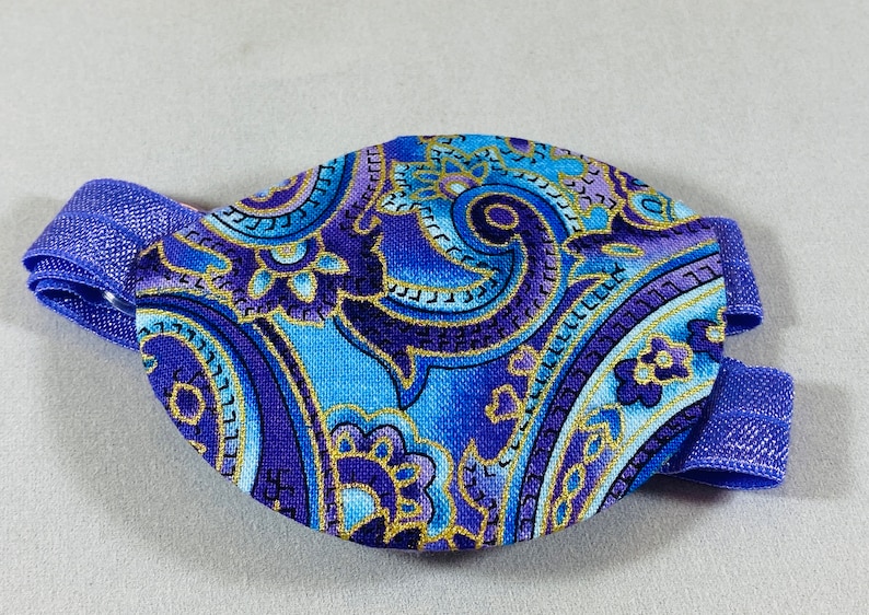 Pode incluir: Um tapa-olho azul e roxo com um padr&atilde;o paisley e detalhes dourados. O tapa-olho est&aacute; preso a uma faixa el&aacute;stica azul.
