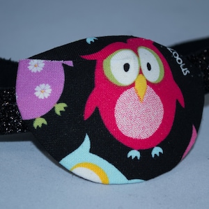 Op de afbeelding: Een zwart ooglapje met een roze uil en andere kleurrijke ontwerpen op een zwarte achtergrond. Het ooglapje heeft een zwart elastisch bandje met glitter.