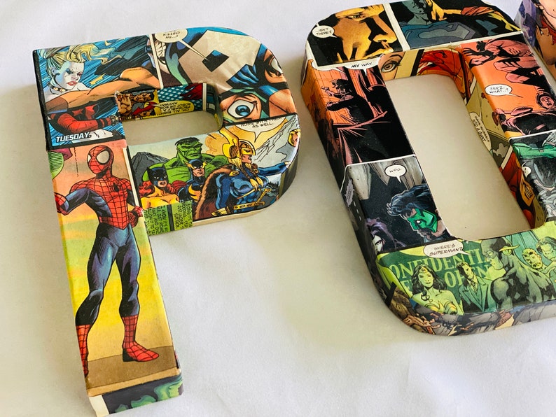 Op de afbeelding: Twee grote kartonnen letters, "P" en "D", bedekt met kleurrijke comicboekpagina's met superhelden zoals Spider-Man, Hulk en Captain America. De letters staan rechtop op een wit oppervlak.