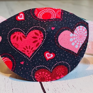 May include: A black eye patch with a pink elastic band. The eye patch is decorated with a repeating pattern of red and pink hearts on a black background.
