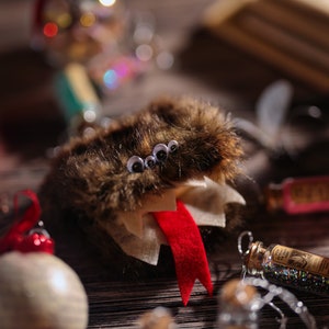 Puede incluir: Un adorno de Navidad de color marr&oacute;n y blanco peludo con una lengua roja y cuatro ojos saltones. El adorno est&aacute; colgado de una cuerda y est&aacute; rodeado de otras decoraciones navide&ntilde;as.