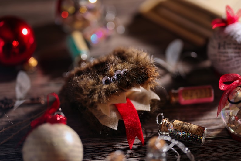 Puede incluir: Un adorno de Navidad de color marr&oacute;n y blanco peludo con una lengua roja y cuatro ojos saltones. El adorno est&aacute; colgado de una cuerda y est&aacute; rodeado de otras decoraciones navide&ntilde;as.