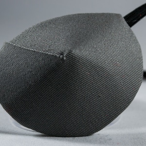May include: A gray eye patch with a black elastic strap. The patch is made of a soft, textured fabric.