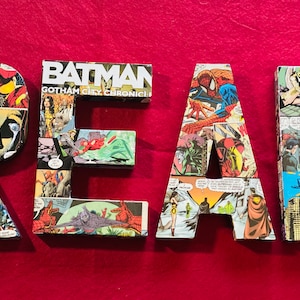 Op de afbeelding: Vier grote letters die "READ" spellen, gemaakt van comicboekpagina's. De letters zijn in verschillende kleuren en bevatten afbeeldingen van superhelden zoals Batman, Superman en Spiderman.