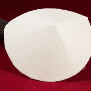 May include: A white eye patch with a rounded shape and a dark brown elastic band. The eye patch is made of a textured fabric and is designed to cover the eye. The background is a deep red color.