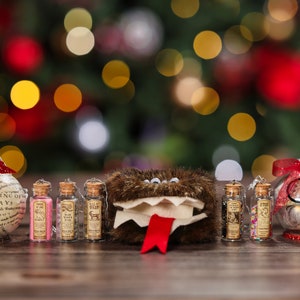 May include: A set of Christmas ornaments featuring a brown furry monster, a glass ornament with the text "The Parting of the Ways", and three small glass bottles with cork tops. The bottles contain glitter and are labeled with different names. The ornaments are arranged on a wooden surface with a blurred background of a Christmas tree with lights.