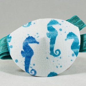 May include: A white eye patch with a teal blue elastic strap. The patch features a repeating pattern of blue seahorse silhouettes on a white background with teal blue watercolor splatter accents.