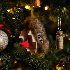 Puede incluir: Un &aacute;rbol de Navidad decorado con adornos, incluyendo un adorno de piel marr&oacute;n con ojos saltones, un adorno de botella de vidrio con tap&oacute;n de corcho y un adorno blanco con purpurina roja y la letra "H" en &eacute;l.
