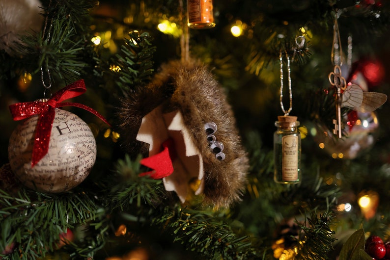 Puede incluir: Un &aacute;rbol de Navidad decorado con adornos, incluyendo un adorno de piel marr&oacute;n con ojos saltones, un adorno de botella de vidrio con tap&oacute;n de corcho y un adorno blanco con purpurina roja y la letra "H" en &eacute;l.