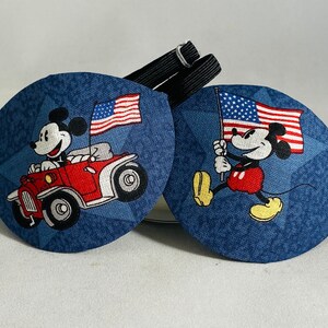 May include: Two blue eye patches with a Mickey Mouse design. One patch features Mickey driving a red car with an American flag. The other patch features Mickey holding an American flag.