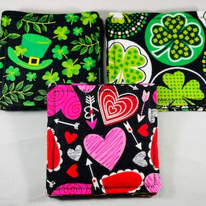 May include: Three fabric coasters with different designs. The first coaster has a black background with green shamrocks and a green leprechaun hat. The second coaster has a black background with green shamrocks and white polka dots. The third coaster has a black background with pink and red hearts and arrows.
