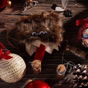 Puede incluir: Una colecci&oacute;n de adornos navide&ntilde;os con tem&aacute;tica de Harry Potter. Los adornos incluyen una criatura peluda marr&oacute;n con ojos saltones, un adorno de vidrio transparente con un par de gafas y un lazo rojo, y un adorno blanco con el texto "The Parting of the Ways".