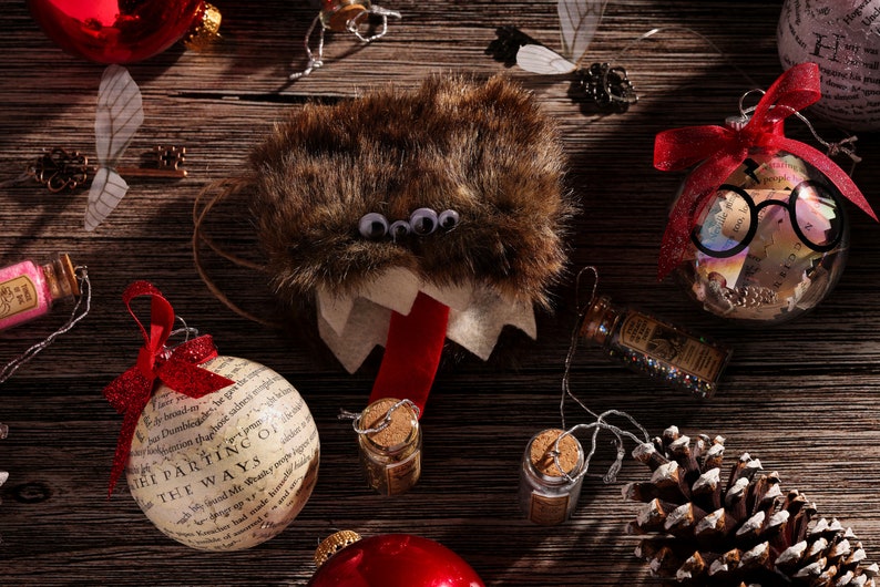 Puede incluir: Una colecci&oacute;n de adornos navide&ntilde;os con tem&aacute;tica de Harry Potter. Los adornos incluyen una criatura peluda marr&oacute;n con ojos saltones, un adorno de vidrio transparente con un par de gafas y un lazo rojo, y un adorno blanco con el texto "The Parting of the Ways".