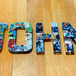 Op de afbeelding: De naam "JOHN" is gespeld in grote, kleurrijke letters. Elke letter is gemaakt van een collage van comicboekpagina's met superhelden en schurken. De letters zijn gerangschikt op een houten oppervlak.