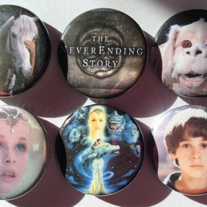 May include: Six round buttons featuring characters and scenes from the movie The NeverEnding Story. The buttons feature a young girl with a white horse, a title card with the movie title, a furry creature with large eyes, a young girl with a crown, a scene with a young boy on a white horse flying through the air, and a young boy with brown hair.