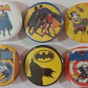 May include: A collection of six round Batman-themed pinback buttons. The buttons feature various comic book illustrations of Batman, Robin, and other characters, with bold colors and the Batman logo.