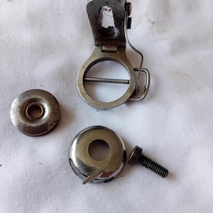 May include: A collection of silver-toned metal sewing machine components including a circular bobbin case with a hinged latch, a separate metal bobbin cover plate with a central hole, and a small black threaded screw for assembly and attachment.