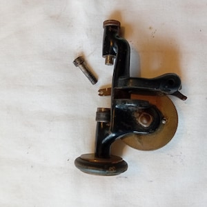 May include: An antique black metal tool with a circular brass blade and a detached bolt. The tool has a curved handle and a base with a rounded bottom. The brass blade has a serrated edge.