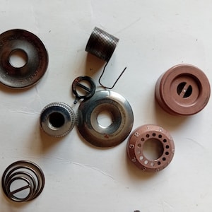 May include: A selection of vintage sewing machine components, including metal bobbins, a spring, and a circular piece with the word "SINGER" engraved. The parts are in shades of brown and silver, arranged on a light surface.