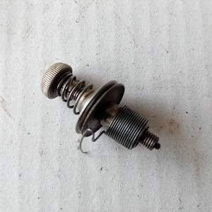 Puede incluir: Primer plano de un conjunto de resorte metálico con un pomo moleteado, un resorte helicoidal y una sección roscada. El conjunto está hecho de componentes metálicos plateados y oscuros, probablemente para un dispositivo mecánico.