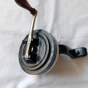 May include: A vintage manual hand crank mechanism featuring a circular black metal housing, a silver-toned metal arm, and a dark brown wooden handle. The device includes a black mounting bracket extending to the right, secured with a visible screw.