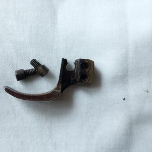 Può includere: Un piedino per macchina da cucire vintage e due viti, probabilmente per riparazione o restauro. Il piedino è un pezzo di metallo curvo con una finitura scura e invecchiata. Le viti sono piccole e metalliche, con segni di usura.