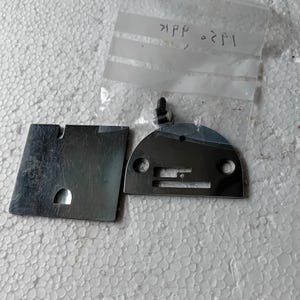 May include: A set of sewing machine parts, including a silver-colored needle plate and a silver-colored presser foot. A screw is attached to the presser foot. The parts are on a white, textured surface.