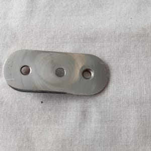 May include: A flat, elongated, silver-coloured metal plate with rounded ends. The plate features three circular holes aligned horizontally along its centre, with a smooth, reflective surface showing subtle circular polishing marks across the metal.