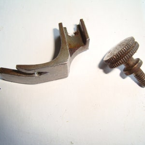 Puede incluir: Un pie de máquina de coser plateado con un brazo de metal curvo y un pequeño tornillo redondo roscado con una cabeza moleteada.