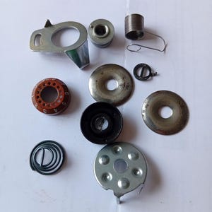 Puede incluir: Un conjunto de piezas de máquina de coser, que incluye canillas metálicas, muelles y varios componentes circulares. Las piezas son de color plateado, marrón y negro, dispuestas sobre una superficie blanca.