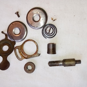 Op de afbeelding: Een assortiment vintage metalen machineonderdelen, waaronder ronde platen, een veer, een draadstang en een kleine schroef. De onderdelen zijn in verschillende tinten bruin en zilver, verspreid op een wit oppervlak.