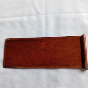 May include: A rectangular wooden tray with a smooth, reddish-brown finish and visible wood grain patterns. The tray features a flat base and a single raised, angled edge on the right side, providing a simple and functional design for holding small items.