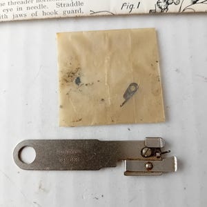 May include: A vintage sewing machine needle threader, a metal tool with a loop, a rectangular body, and a small attached mechanism. The text "121632 SIMANCO" is visible. A small, folded paper packet and a portion of a paper with text are also present.