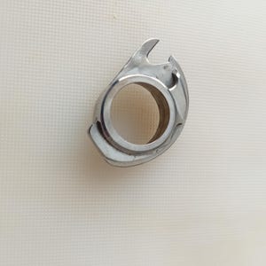 Könnte beinhalten: Eine silberfarbene Nähmaschinen-Spulenkapsel aus Metall mit einer kreisförmigen Mittelöffnung, einem geschwungenen Außenrand und einem kleinen spitzen Vorsprung an der Oberseite. Das Bauteil hat eine glatte, polierte Oberfläche und eine komplexe Form.