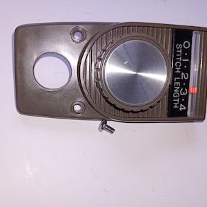 May include: A brown sewing machine stitch length control panel with a dial and a scale marked 0-4. The panel has a circular opening and two screw holes. A screw is visible near the panel.