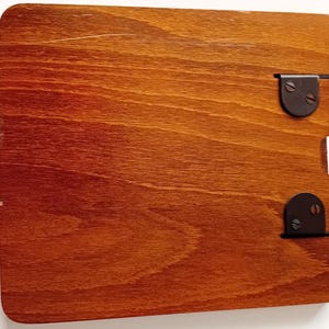 May include: A rectangular, stained wooden board with rounded corners. Two black metal hinges are attached, one at the top and one at the bottom. The wood has a warm, reddish-brown hue.