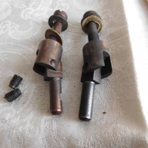 Może przedstawiać: Dwie metalowe części z gwintowanym trzpieniem i kwadratową głowicą. Części są w różnych kolorach, jedna jest miedziana, a druga czarna. Na białej powierzchni znajdują się dwa małe śruby.