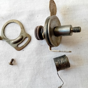 May include: A disassembled metal sewing machine tension assembly featuring a central cylindrical post with a threaded end, a circular tension disc, a small knurled nut, a separate metal plate with a slot, a small screw, and a coiled metal spring.