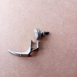 May include: A silver sewing machine presser foot, featuring a curved toe and a screw fitting. This component is designed to secure fabric during sewing. The image is set against a tan backdrop.