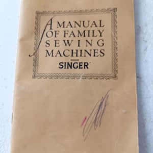 Könnte beinhalten: Ein Vintage-Handbuch mit dem Titel "A Manual of Family Sewing Machines" von Singer. Das Cover ist in einem verblichenen Beigeton gehalten und hat schwarzen Text und dekorative Bordüren. Das Handbuch hat einige Markierungen auf dem Cover.