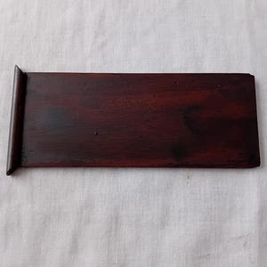 May include: A rectangular, dark brown wooden board with a smooth, polished finish. The board features a distinct, raised vertical edge on the left side, creating a small lip. The wood grain is visible across the surface, showing deep mahogany tones.