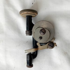 May include: An antique hand-cranked drill with a wooden handle and a metal body. The drill features a circular saw blade and a metal clamp. The drill is black with silver and brown accents.