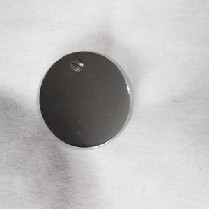 Puede incluir: Una tapa redonda de metal plateado con un pequeño tornillo en el centro.