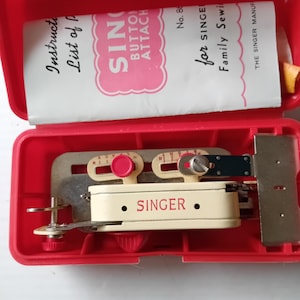 May include: A red plastic Singer button attachment kit, open to reveal the sewing machine accessory. The kit includes a cream-colored attachment with the Singer logo, a metal plate, and a booklet. The booklet reads "Singer Button Attach".