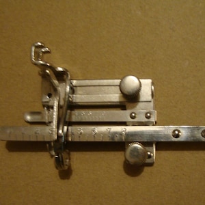 May include: A silver sewing machine attachment with a ruler, two knobs, and a metal hook. The ruler has markings from 1 to 10.