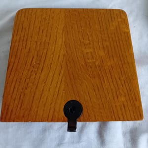 May include: A square wooden box with a warm, medium-brown wood grain finish. The lid features a smooth, flat surface with rounded corners. A small, circular black metal latch is centred on the front edge, providing a secure closure mechanism.