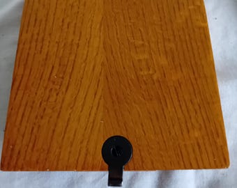 May include: A square wooden box with a warm, medium-brown wood grain finish. The lid features a smooth, flat surface with rounded corners. A small, circular black metal latch is centered on the front edge, providing a secure closure mechanism.