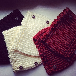 Puede incluir: Tres calienta piernas de bebé de ganchillo en negro, blanco y rojo. Cada calienta piernas tiene un cierre de botón.