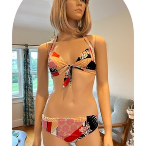 May include: A two-piece bikini set with a halter top and low-rise bottoms. The bikini features a colorful geometric pattern in shades of pink, red, black, and beige. The top ties in the front, and the bottoms have a matching design. The bikini is displayed on a mannequin.