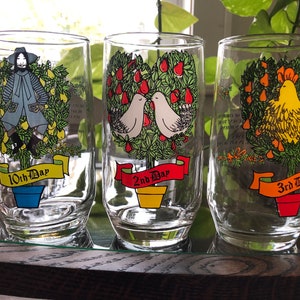 May include: Set of six clear glass tumblers with colorful illustrations of a pear tree, a rooster, and a man. Each glass has a red ribbon with the words "10th Day", "2nd Day", and "3rd Day" printed on it.